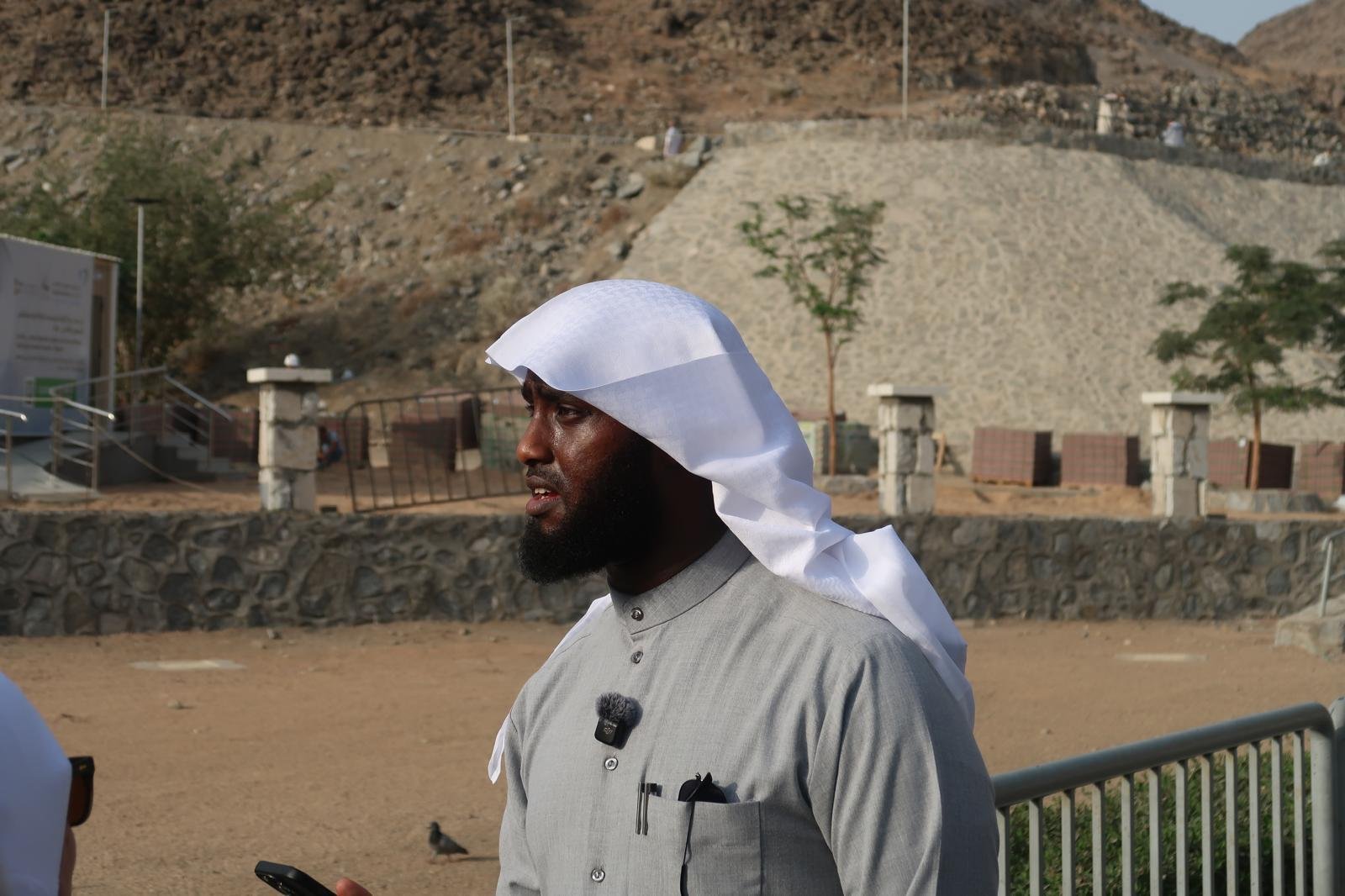 A man wearing a white headscarf and gray traditional attire standing outdoors in a desert-like area with rocky terrain and sparse trees, holding a smartphone and wearing a microphone clipped to his shirt.
