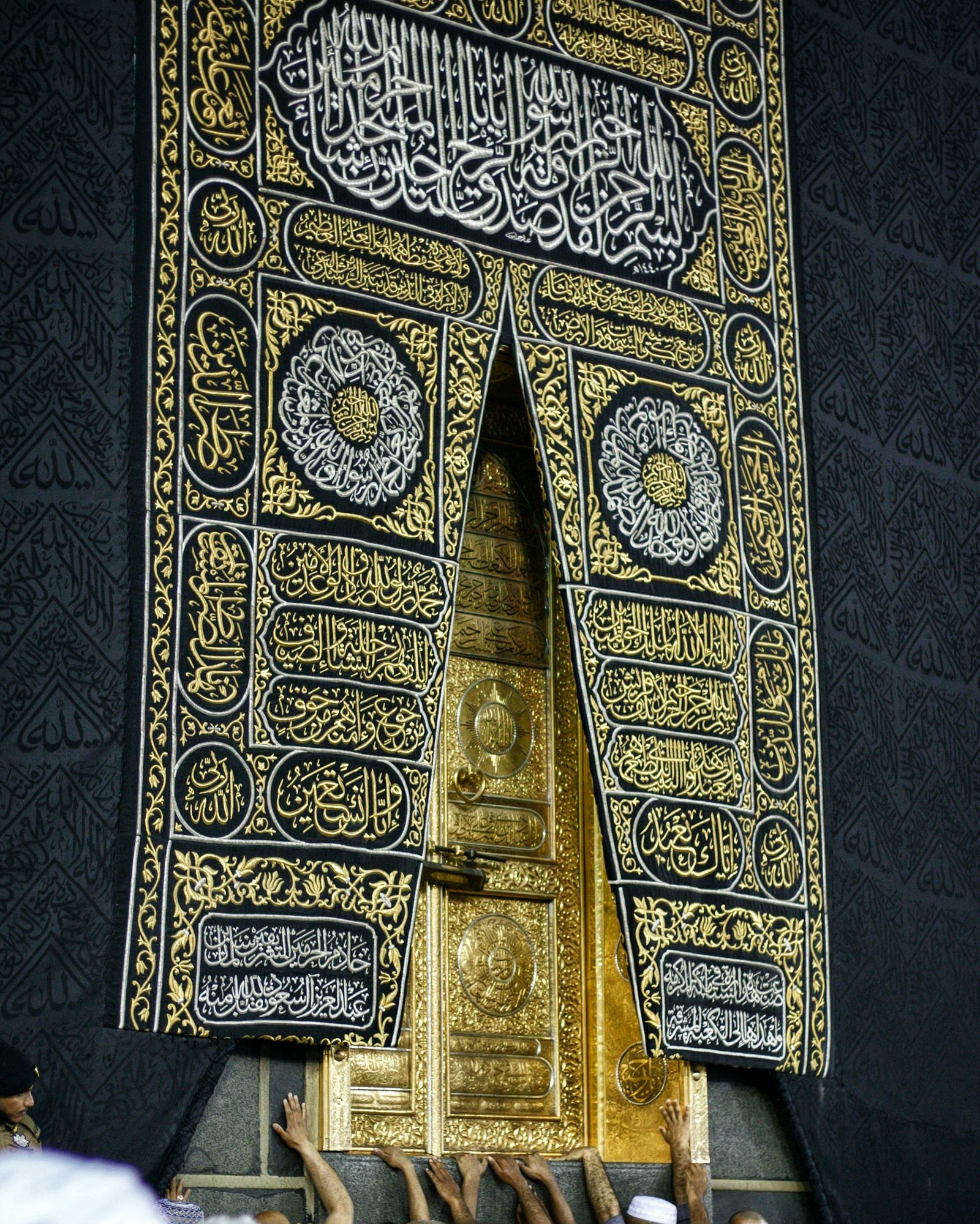 Close-up of the ornate gold door of the Kaaba in Mecca, partially covered by the black and gold-embroidered Kiswah cloth with Arabic calligraphy, with several hands reaching to touch the door from below.