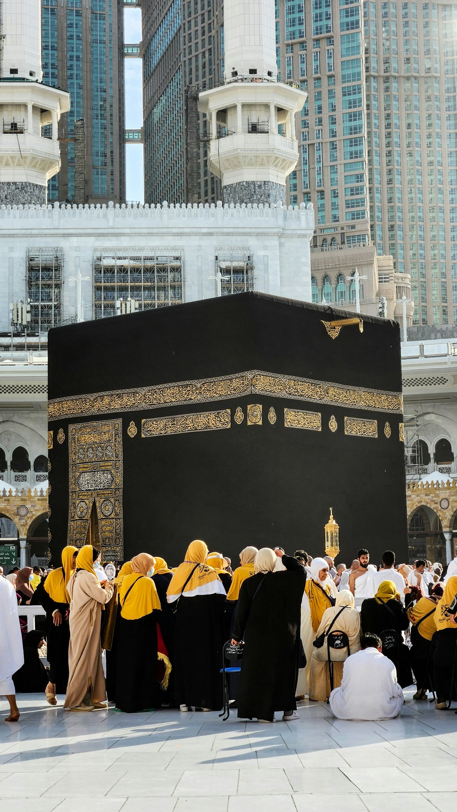 Group of pilgrims, many women wearing yellow headscarves, gathered near the Kaaba in the Masjid al-Haram mosque, Mecca, with tall buildings in the background.